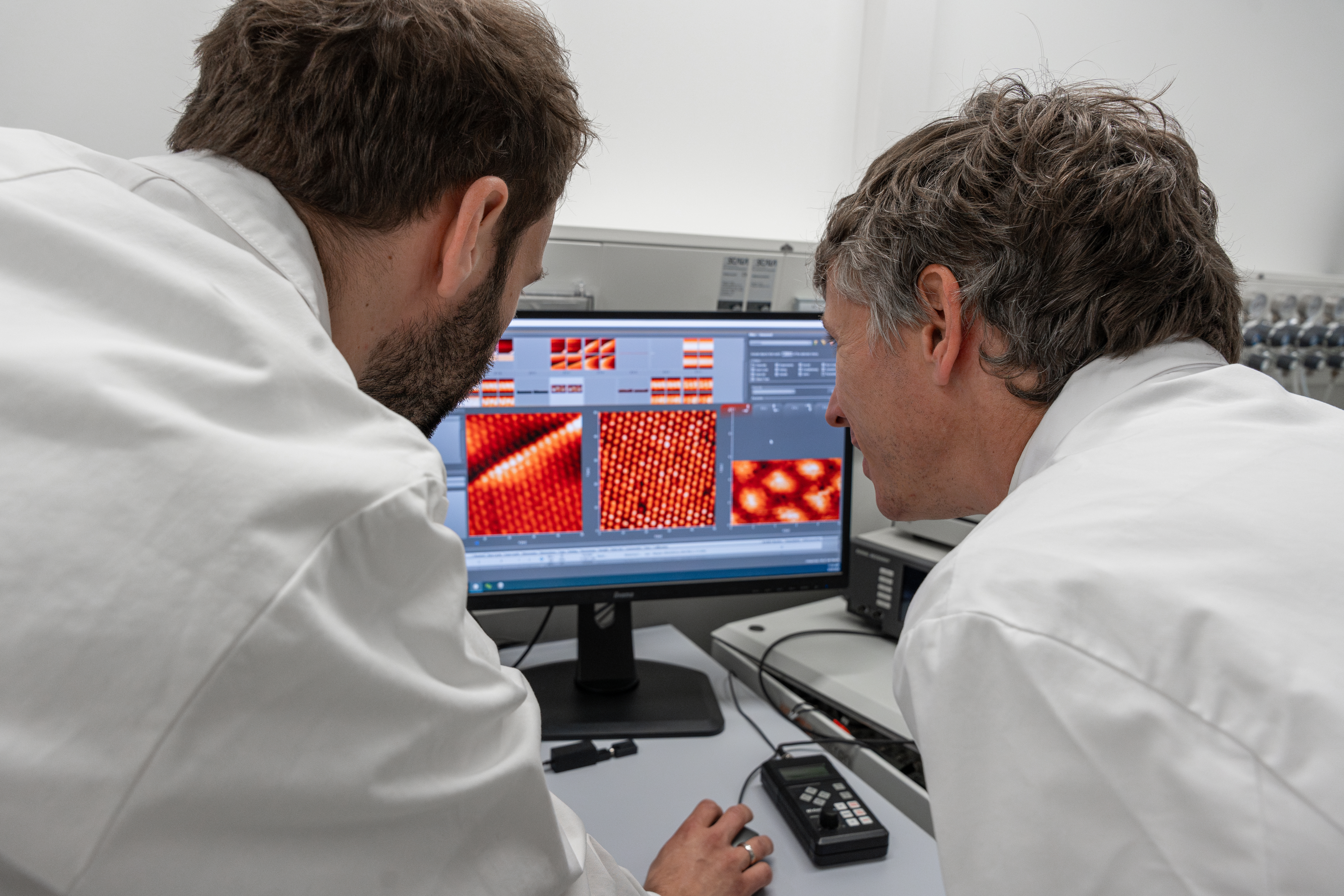Zwei Männer in weißen Forschungskitteln sind von hinten zu sehen, wie sie auf einen Computerbildschirm schauen. Auf diesem sind Mikroskopaufnahmen von optischen 2D Materialien zu sehen.