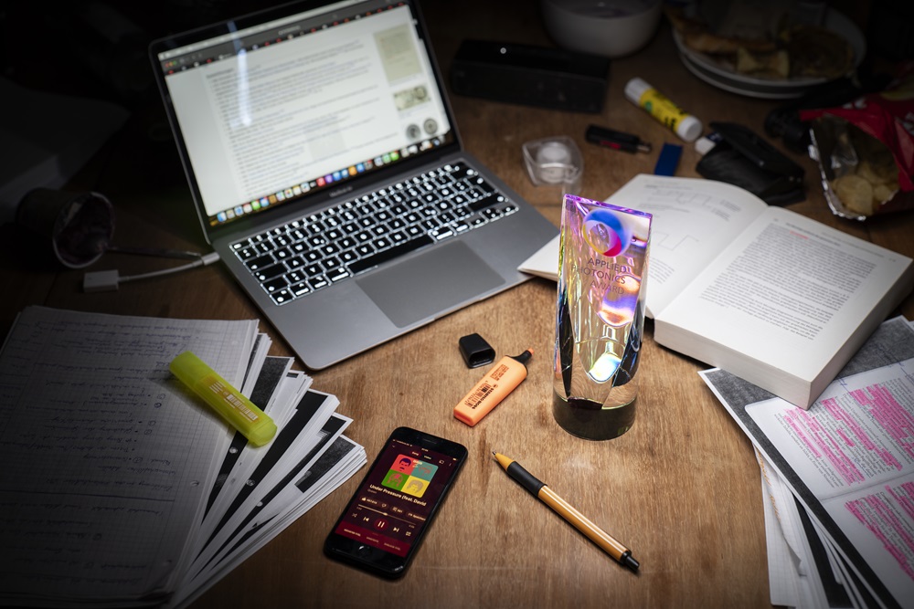 A glass trohpy  stands on a desk, surrounded by work materials such as books, pens, a computer and a smartphone.