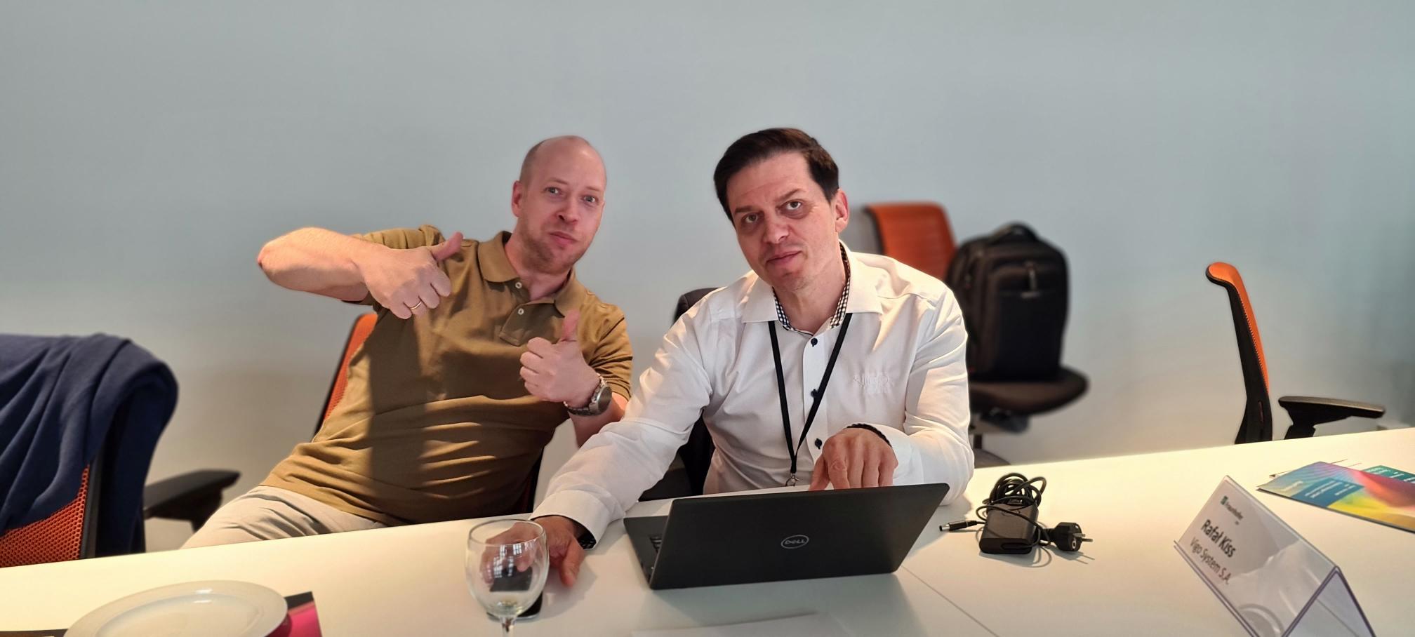 Two men sit together at the computer and give the thumbs up to the camera at the Photonics Manager Compact 2022.